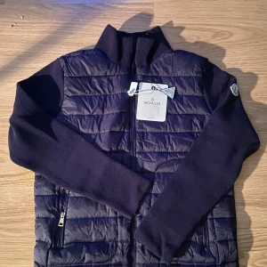 Mörkblå quiltad cardigan jacka Moncler - Snygg mörkblå cardigan jacka från Moncler med quiltad framsida och stickade ärmar. Jackan har dragkedja framtill, hög krage och Moncler-logga på vänster ärm. Perfekt för dig som vill ha en stilren och exklusiv look.