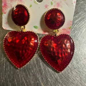 Röda hjärtformade örhängen i 18k guldpläterad  - Säljer ett par statement örhängen med stora röda hjärtan och rund topp i metallic-look. Örhängena har glittrig yta och guldfärgade kanter som ger extra bling. Perfekt för dig som vill sticka ut med färg och cool form. Fästs med klassisk stift.I 18k guldpläterad.