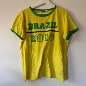 Gul Brazil 2018 t-shirt med gröna detaljer - Gul t-shirt med gröna kanter och tryck 'BRAZIL 2018' på bröstet. Rund halsringning och korta ärmar med gröna muddar. Perfekt för dig som gillar fotboll och vill visa support för Brasilien. Skön och lätt i bomull.