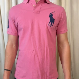 Rosa pikétröja från Polo Ralph Lauren - Snygg rosa pikétröja från Polo Ralph Lauren i custom fit. Tröjan har korta ärmar, klassisk krage och stor marinblå logga på bröstet samt en broderad siffra 3 på ena ärmen. Tillverkad i mjuk bomull som är skön mot huden.