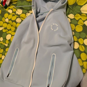Ljusblå hoodie från BOSS med dragkedja - Snygg ljusblå hoodie från BOSS med vit dragkedja och vita detaljer runt fickorna. Tröjan har huva med snörning och BOSS-logga tryckt på bröstet. Perfekt för dig som gillar stilrena och sportiga plagg.