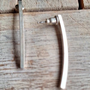 Silvriga minimalistiska örhängen - Säljer ett par stilrena örhängen i silverfärgad metall med rak och lätt böjd form. De är cirka 4 cm långa och har en enkel, modern design som passar dig som gillar minimalistiska smycken. Perfekta för att ge din look en clean touch.
