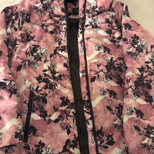 Blommig rosa vindjacka från Everest - Säljer en snygg vindjacka från Everest med rosa och vita toner, blommigt mönster och svarta detaljer. Jackan har dragkedja framtill och praktiska fickor. Perfekt för dig som gillar färg och mönster. Märke syns på bröstet. Överdrag vuxen model passar som s jätte fin varm 