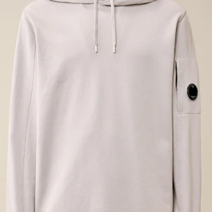 Ljusgrå hoodie från C.P. Company - Snygg ljusgrå hoodie från C.P. Company med klassisk linsdetalj på ärmen. Tröjan har huva med snörning och långa ärmar med ribbade muddar. Tillverkad i mjuk bomull som är skön mot huden. Perfekt för dig som gillar stilrena och moderna plagg. Skriv om ni är intresserade eller vill ha mer bilder finns tillgänglig varje dag🙌