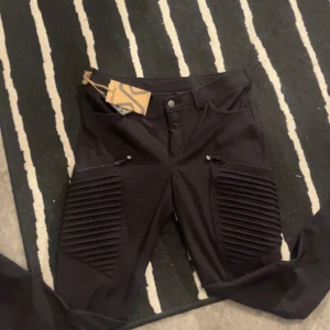 Svarta RVRC Pusher jeans - Svarta jeans från RVRC med ribbade paneler på låren och dragkedjor vid fickorna. Snygg, modern look med normal passform och midwaist. Materialet är slitstarkt och passar perfekt för dig som gillar streetstyle och funktionella detaljer.