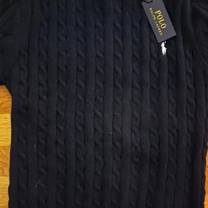 Svart kabelstickad tröja Polo Ralph Lauren - Svart kabelstickad tröja från Polo Ralph Lauren med rund hals och långa ärmar. Tröjan har klassisk ribbad kant vid halsen och nederdelen, och är tillverkad i mjukt material som känns lyxigt. Perfekt för dig som gillar stilrena och tidlösa plagg.