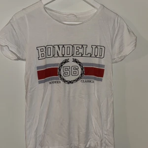 Vit Bondelid t-shirt med tryck XS - Vit t-shirt från Bondelid i storlek XS med coolt tryck framtill, röd och grå rand samt texten 'Modern Classics 56'. Rund hals och korta ärmar, tillverkad i mjuk bomull. Perfekt för dig som gillar sportig och avslappnad stil.
