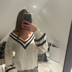 Vit kabelstickad v-ringad tröja - Vit kabelstickad tröja med v-ringning och breda muddar. Tröjan har mörkblå och vinröda ränder vid halsringning och ärmslut. Den har en loose passform och är perfekt för lager-på-lager-stil under höst och vinter.