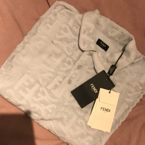 Vit Fendi piké  med logomönster - Säljer en vit piké från Fendi i storlek S med ikoniskt Fendi-logomönster över hela plagget. Piké  har klassisk krage, korta ärmar och knappar framtill. Materialet är mjukt och frottéliknande, vilket ger en unik och lyxig känsla. Perfekt för dig som gillar designerplagg med tydlig branding.