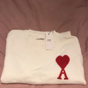 Vit stickad tröja med rött hjärta från Ami - Vit stickad tröja från Ami med rund hals och ribbade muddar. På bröstet finns ett stort rött hjärta med bokstaven A under, vilket ger en unik och snygg detalj. Perfekt för dig som gillar stilrena plagg med en twist.