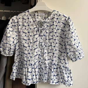Vit blommig blus - 🤍Säljer en vit blus från Holly & Whyte med blått blommigt broderat mönster. Blusen har snörning fram, puffiga korta ärmar och en volang nedtill 🤍