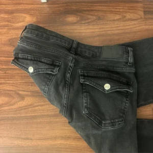 Svarta jeans med ficklock - Snygga svarta jeans med coola ficklock och silverfärgade knappar på bakfickorna. Jeansen har en klassisk femficksdesign och är tillverkade i ett mjukt denimtyg som sitter skönt. Perfekta för dig som gillar en stilren look med extra detaljer.