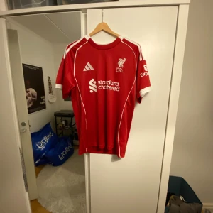 Liverpool röd fotbollströja Adidas - Säljer en röd Liverpool fotbollströja från Adidas med vita detaljer och tryck. Fram har tröjan Liverpool FC-logga och sponsortryck, bak står ISAK och nummer 9 i vitt. Klassisk rund hals och korta ärmar. Perfekt för dig som älskar Premier League och vill sticka ut på läktaren.