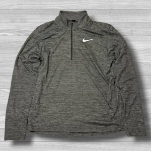 Grå Nike långärmad träningströja - Nike långärmad träningströja i melerad grå färg med halv dragkedja och hög krage. Swoosh-logga på bröstet och meshfoder på insidan för extra ventilation. Perfekt för löpning eller gym. Snygg och sportig stil. Axlar->Midja = 74,5cm. Vänster -> Höger = 50,5 cm