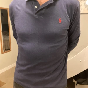 Mörkblå pikétröja från Polo Ralph Lauren - Säljer en mörkblå långärmad pikétröja från Polo Ralph Lauren i custom slim fit. Tröjan har klassisk krage, knäppning vid halsen och den ikoniska röda broderade loggan på bröstet. Tillverkad i mjuk bomull som är skön mot huden. Tröjan är inte använd och är i mkt bra skick, pris kan diskuteras vid snabb affär.  Obs!! Moddelen på bilden har en större storlek i vanliga fall vilket gör att tröjan ser lite liten ut…