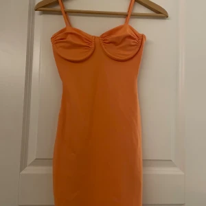 Orange kort klänning med kupor - Snygg orange kort klänning med smala axelband och markerade kupor framtill. Klänningen har en figurnära passform och är perfekt för dig som gillar färgstarka plagg med enkel design. Materialet känns mjukt och stretchigt mot kroppen.