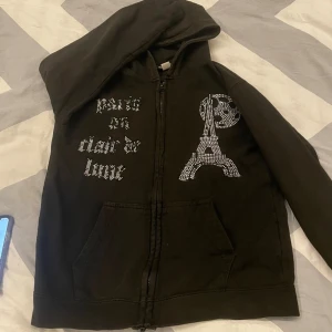 Blinglucid zip. - Svart hoodie från Blinglucid med dragkedja, huva och fickor. Framsidan har coolt strassmönster med Eiffeltornet och texten 'Paris au clair de lune'. Tillverkad i mjuk bomull och polyester, perfekt för dig som gillar statement-plagg. Märket går inte heller att köpa längre för företaget har lagt ner. Jätte bra för din son i julklapp/present. Storlek xs.