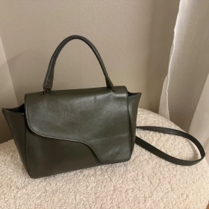 Mörkgrön handväska i skinn från ATP atelier  - Snygg mörkgrön handväska med stilren design och asymmetrisk lock. Väskan har både handtag och axelrem, samt guldfärgade metalldetaljer. Tillverkad i slätt skinn och passar perfekt för dig som gillar minimalistisk stil. Använt skick men mycket kvar att ge 