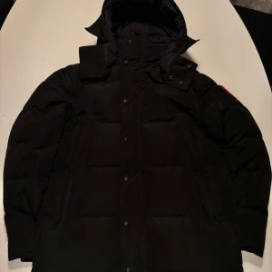 Svart dunjacka från Canada Goose - Säljer en svart dunjacka från Canada Goose med klassisk patch på ärmen. Jackan har huva, stora fickor och stängs med både dragkedja och knappar. Perfekt för kalla dagar och har en clean, oversized look som är riktigt nice. Det finns en ända defekt ,ed jackan och det är att en av knapparna har gått sönder men det syns inte! Annars såhär det en stilren jacka som är perfekt för vintern ❄️ **PRIS KAN DISKUTERAS**