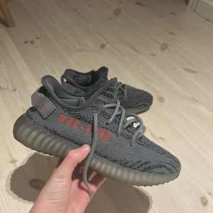 Adidas Yeezy Boost 350 V2 grå - Säljer ett par Adidas Yeezy Boost 350 V2 i mörkgrå färg med stickad ovandel och tydliga orange detaljer på sidan. Skorna har snörning, platt sula och rund tå. Den stickade strukturen ger en unik look och skön passform. Perfekt för dig som gillar streetwear och sneakers med hype.