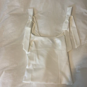 Vit linnetopp med rosettband - vit linnetopp med breda axelband med stora rosetter. Toppen har en rak och croppad passform 