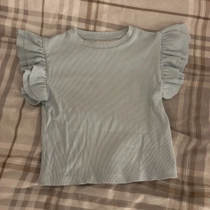 Ljusblå ribbad topp med volangärmar - Supersöt ljusblå topp med ribbad struktur och stora volangärmar. Toppen har rund halsringning och är tillverkad i ett mjukt, stretchigt material som känns skönt mot huden. Perfekt för dig som vill ha en söt och trendig look.