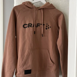 Beige hoodie från Craft XS - Säljer en beige hoodie från Craft i storlek XS. Tröjan har huva med snörning, stor magficka och svart logga framtill. Mjuk insida och långa ärmar, perfekt för chill eller träning. Klassisk och enkel design som funkar till det mesta.