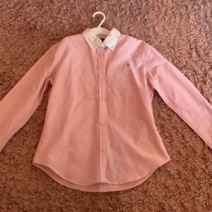 Rosa skjorta från Ralph Lauren XL - Snygg rosa skjorta från Ralph Lauren i storlek XL med vit klassisk krage och knappar framtill. Tillverkad i 100% bomull, långärmad och med en stilren, normal passform. Perfekt för dig som gillar preppy stil och vill ha något som sticker ut lite extra.