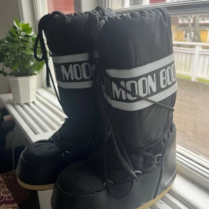 Svarta Moon Boots med snörning - Stlk 39/41. Moon Boots i svart med vit logga och snörning framtill. Höga skaft och platt sula med gult och svart mönster undertill. Tillverkade i syntetmaterial med vadderad insida för extra värme. Perfekta för kalla vinterdagar och snö.
