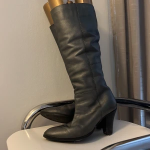 Silvriga höga stövlar med klack - Säljer ett par svarta stövlar i skinn med snygg spetsig tå och stabil klack. Stövlarna är höga och har en stilren, klassisk design som passar till många olika outfits. Perfekt för dig som vill ha en cool och tidlös look.