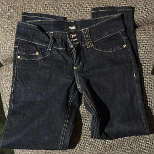 Mörkblå jeans - Snygga mörkblå jeans från heart quate (tror jag det står) med kontrasterande vita sömmar och coola broderade detaljer på bakfickorna. Modellen har låg midja, raka ben och klassisk femficksdesign. Jeansen är tillverkade i slitstarkt denim och har tre knappar och dragkedja framme. Storlek står inte men skulle säga 32 då höfterna är ganska små (W25 typ)