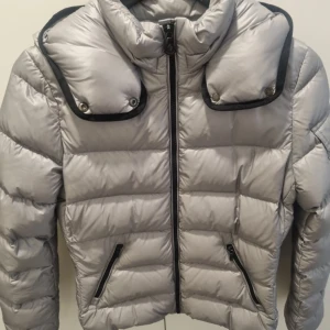 Silver pufferjacka från Moncler - Snygg silverfärgad pufferjacka från Moncler med quiltad design och hög krage. Jackan har svarta detaljer, två fickor med dragkedja och Moncler-logga på ärmen. Perfekt för kalla dagar litet hål på armbågen och lite fläckar kolla bilder❤️