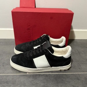 Valentino Garavani svartvita sneakers - Svartvita Valentino Garavani sneakers i mocka och skinn med klassisk låg siluett. Skorna har svarta snören, vit panel på sidan och coola nitar på sulan baktill. Perfekta för dig som vill ha en stilren men edgy look.