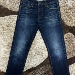 Mörkblå jeans från Jack & Jones 29/30 - Snygga mörkblå jeans från Jack & Jones i storlek 29/30. Modellen har klassisk femficksdesign, raka ben och subtila slitningar framtill för en cool look. Jeansen är tillverkade i slitstarkt denim med gul kontrastsöm och läderpatch bak i midjan.