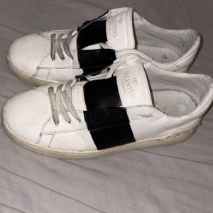 Valentino Garavani Open sneakers vit/svart - Säljer ett par Valentino Garavani Open sneakers i vitt skinn med bred svart rem över mitten. Klassisk rund tå, platt sula och snörning framtill. Snygga nitar längs hälen ger extra edge. Perfekt för dig som gillar lyxiga och stilrena sneakers.