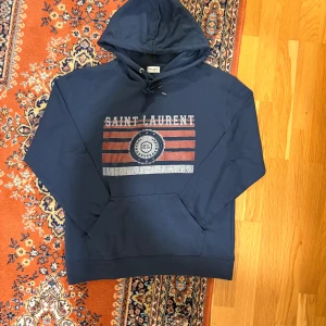 Blå hoodie från Saint Laurent - Snygg marinblå hoodie från Saint Laurent med tryck framtill i rött, vitt och blått. Tröjan har huva med snörning och en stor magficka. Perfekt för dig som gillar streetwear och vill ha något som sticker ut med exklusiv vibe.