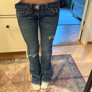 True Religion blå bootcut jeans - Snygga blå jeans från True Religion med bootcut och låg midja. Klassiska fickor med dekorativa sömmar och logga bak. Jag har sytt de lite så de blev tightare men enkelt att sprätta upp (har inte klippt bort något) midjemåttet är 38 cm och innerbenslängden är är ca 77 cm. 