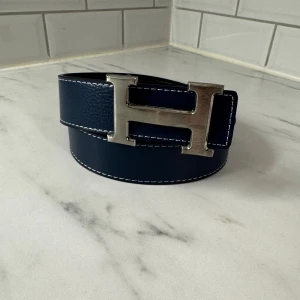 H bälte  med H-spänne marinblå - Säljer ett stilrent H bälte i marinblått och svart skinn med grov struktur. Bältet har ett ikoniskt H-spänne i silverfärgad metall och vit kontrastsöm längs kanterna. Perfekt accessoar för att lyfta din outfit med lyxig känsla och klassisk design.