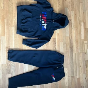 Trapstar svart hoodie med broderi - Svart hoodie från Trapstar med broderad logga i blått, rött och vitt framtill. Tröjan har huva med snörning och en stor magficka. Materialet är mjukt och känns som bomull eller fleece, perfekt för chill dagar. Snygg streetwear-vibe med färgglada detaljer.