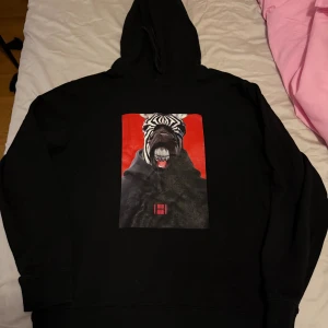 Svart hoodie med zebratryck från Hugo - Svart hoodie från Hugo med stor, färgstark print på bröstet som visar en zebra med cool attityd mot röd bakgrund. Tröjan har huva och ribbade muddar. Perfekt för dig som vill sticka ut med ett unikt motiv och streetwear-vibe.