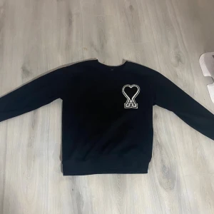 Svart sweatshirt från Ami med hjärtdetalj - Svart sweatshirt från Ami med lång ärm och en stor vit broderad hjärtdetalj på bröstet. Tröjan har en klassisk rund hals och är tillverkad i mjuk bomull som är skön mot huden. Perfekt för dig som gillar stilrena plagg med en unik twist.