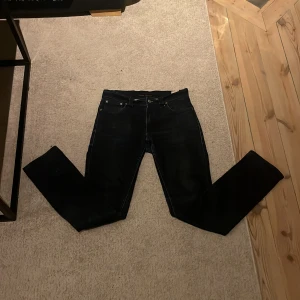 Mörkblå Nudie jeans - Mörkblå jeans från Nudie Jeans med klassisk femficksdesign och orange logopatch bak. Jeansen har thin finn passform, tillverkade i slitstarkt bomullsmaterial. Perfekta för dig som gillar stilrena och tidlösa jeans.