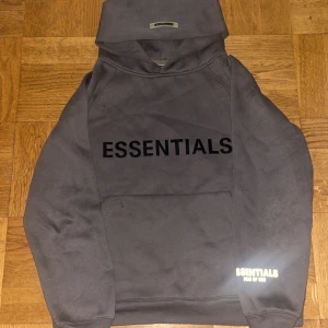 Essentials hoodie  - Säljer en mörkgrå hoodie från Fear of God Essentials med stor svart logga på bröstet och mindre tryck på ärmen. Tröjan har huva, känguruficka och är långärmad. Perfekt för dig som gillar streetwear och vill ha en clean look.