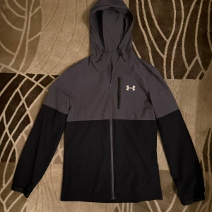 Grå och svart vindjacka Under Armour - Snygg tvåfärgad vindjacka från Under Armour i grått och svart. Jackan har huva med snörning, lång ärm, hel dragkedja och en bröstficka med zip. Använd 1-2 ggr, säljs då den inte riktigt passar. 