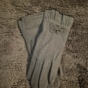 Grå ullhandskar med rosett - Snygga grå handskar i mjuk ull med en liten rosett som detalj vid handleden. Handskarna har en enkel och stilren design med svarta sicksackkanter upptill. Perfekta för dig som vill ha något klassiskt men ändå med en twist.