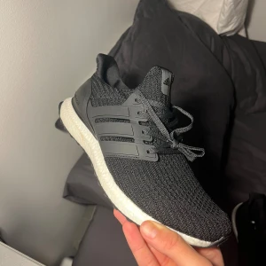 Adidas Ultraboost svarta sneakers - Svarta Adidas Ultraboost sneakers med stickad ovandel och klassiska tre ränder på sidan. Skorna har vit Boost-mellansula och svart yttersula med Torsion System. Perfekta för dig som vill ha både stil och komfort. Snörning framtill och diskret Adidas-logga på plösen.