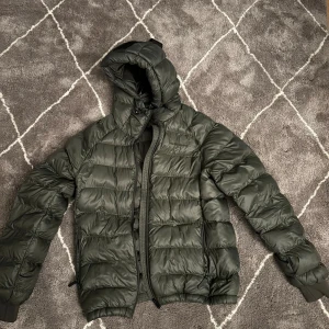 Mörkgrön pufferjacka från Peak Performance - Säljer en mörkgrön pufferjacka från Peak Performance i storlek S. Jackan har huva, dragkedja och ribbade muddar vid ärmslut. Den quiltade designen ger en schysst look och jackan är perfekt när det är kallt ute.