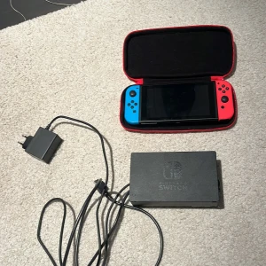 Nintendo Switch - Nintendo Switch med blå och röd Joy-Con, medföljande original dockningsstation, laddare och röd skyddsväska. Konsolen och tillbehören ser ut att vara i gott skick, med några mindre repor på skärmen. Perfekt för både bärbart och stationärt spelande.