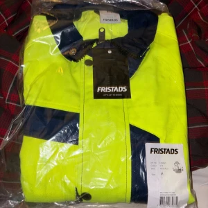Gul och marinblå Fristads vindjacka - Fristads arbetsjacka i varsel med breda reflexband runt midja och ärmar. Jackan har dragkedja, knappar och flera praktiska fickor. Perfekt för dig som vill synas och ha en schysst look på jobbet. Helt oanvänd i förpackning, ny pris 2400, säljer för 500kr