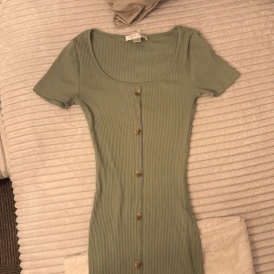 Ribbad grön klänning med knappar - Superfin ribbad klänning i ljusgrönt från FAMIS. Modellen är figurnära med korta ärmar, rundad urringning och snygga knappar längs hela framsidan. Perfekt för dig som gillar en enkel men trendig look.
